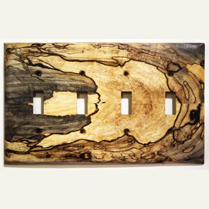 Könnte beinhalten: Eine Holz-Lichtschalterplatte mit drei rechteckigen Öffnungen für Schalter. Die Platte weist ein natürliches Holzmaserungsmuster mit Braun-, Schwarz- und Beigetönen auf. Die Holzmaserung hat ein einzigartiges, wirbelndes Muster.