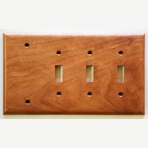 Peut inclure: Une plaque d'interrupteur en bois avec trois ouvertures pour interrupteurs à bascule et six trous de vis. La plaque est en bois brun clair avec un motif de grain de bois visible. Les bords sont légèrement biseautés.