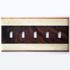 Puede incluir: Una placa de interruptor de luz de madera con cinco aberturas. La placa presenta una sección central marrón oscuro con un borde y detalles de madera más clara. La placa es rectangular y tiene orificios para tornillos visibles.