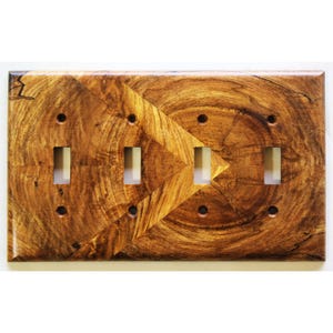 Könnte beinhalten: Eine rechteckige Lichtschalter-Wandplatte aus Holz mit einer natürlichen Holzmaserung, dunkelbraunen Wirbeln und einem chevronartigen Muster. Die Platte hat vier rechteckige Ausschnitte für Schalter und acht kleine Schraublöcher.