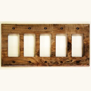 May include: A wooden light switch plate with five rectangular openings. The plate is made of a light brown wood with visible grain and knots. The openings are for the light switches, and the plate is secured with dark screws.