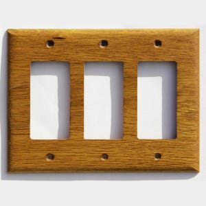 Könnte beinhalten: Eine dreifache Lichtschalterabdeckung aus Holz mit warmem, goldbraunem Finish. Die Abdeckung hat drei rechteckige Öffnungen und sechs Schraubenlöcher. Die Holzmaserung ist sichtbar und verleiht dem Design Textur.