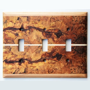 May include: A wooden light switch plate with three switch openings. The plate features a natural wood grain pattern in shades of brown, tan, and black. A thin, light-colored stripe runs horizontally across the center. The frame is a lighter wood.