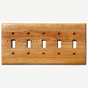 May include: A rectangular, wooden light switch cover with five toggle switch openings. The cover is made of light-colored wood with a prominent grain pattern. There are small screw holes above and below each switch opening.