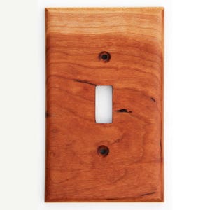 Peut inclure: Plaque d'interrupteur en bois rectangulaire, avec une ouverture pour un interrupteur à bascule et deux trous de vis. Le bois brun rougeâtre présente un grain visible et une finition lisse. Le bois a des stries et des nœuds plus foncés.