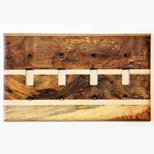 Könnte beinhalten: Eine Holz-Lichtschalterplatte mit vier Öffnungen für Lichtschalter. Die Platte ist aus dunklem Holz mit einer Einlage aus hellem Holz. Das Holz hat eine natürliche Maserung.