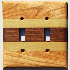 May include: A wooden double light switch cover with a layered design. The top and bottom sections are a light brown with wood grain, separated by a light yellow and a dark brown section in the middle. Four small holes are visible.