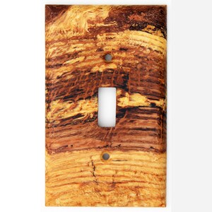 May include: A decorative light switch cover with a wood grain pattern. The cover features a single toggle switch opening and two screw holes. The wood grain design has shades of brown, tan, and black.