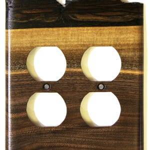 May include: A wooden electrical socket cover with four white outlet holes. The cover has a natural wood grain pattern in shades of brown and tan, with a dark, textured top edge.