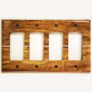 Puede incluir: Una placa de interruptor de luz de madera con cuatro aberturas rectangulares para interruptores. La placa está hecha de madera teñida con un patrón de vetas visible y está asegurada con tornillos oscuros. Una fina franja de color claro recorre horizontalmente la placa.