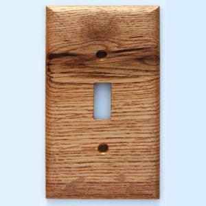 Puede incluir: Una cubierta de interruptor de luz de madera con una sola abertura de palanca. La cubierta es de madera marrón claro con un patrón de vetas visible. Tiene dos pequeños agujeros para tornillos y una abertura rectangular para el interruptor.