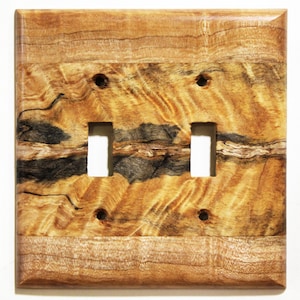 May include: A wooden double light switch cover with a natural wood grain pattern. The cover has two rectangular switch openings and four screw holes. The wood features a mix of light brown, dark brown, and black tones.