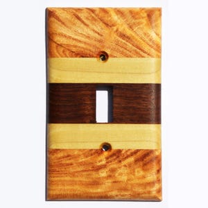 May include: A wooden light switch cover with a striped design. The cover is made of three different types of wood: a light wood, a dark wood, and a reddish-brown wood. The cover has a single switch opening.