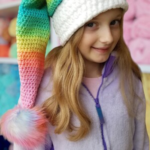 May include: A girl wearing a white crocheted hat with a long rainbow crocheted tail. The hat has a fluffy pom-pom on the end of the tail.