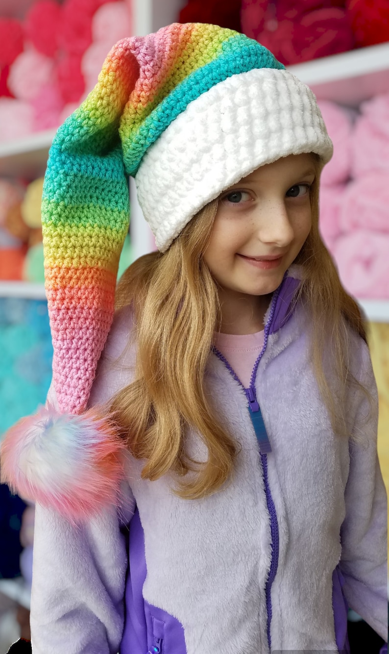 May include: A girl wearing a white crocheted hat with a long rainbow crocheted tail. The hat has a fluffy pom-pom on the end of the tail.
