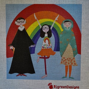 May include: Three women stand in front of a rainbow. The woman on the left is wearing a black robe and glasses. The woman in the middle is wearing a rainbow dress and is holding a rainbow stick. The woman on the right is wearing a green and yellow dress and a headscarf.