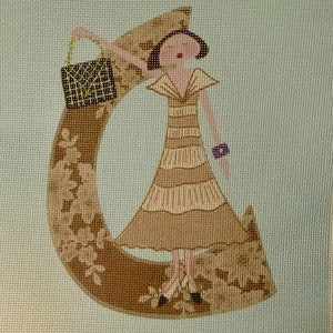 Puede incluir: Un diseño bordado en marrón y beige de una mujer sosteniendo un bolso negro. La mujer está de pie sobre una gran letra C.