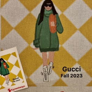 May include: A green sweater with a turtleneck and a large orange bag. The person is wearing sunglasses and white boots. The background is a yellow and white diamond pattern. The text "Gucci Fall 2023" is at the bottom of the image.