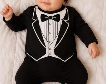 Puede incluir: Un bebé con un body negro estilo esmoquin con una camisa blanca, pajarita y botones. El conjunto incluye pantalones con pies a juego y un gorro negro. El bebé está acostado sobre una superficie blanca.