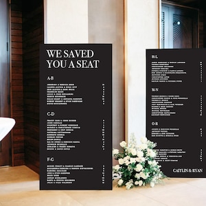Puede incluir: Dos letreros negros con texto blanco que enumeran nombres en orden alfabético. Los letreros están titulados "We Saved You A Seat" y "H-L, M-N, O-R". Los letreros están en un piso con una mesa blanca y una planta en maceta con flores blancas.