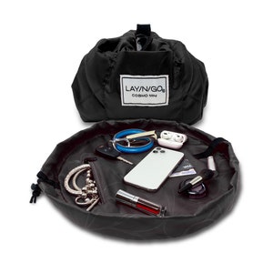 May include: A black lay-n-go cosmo mini makeup bag with a drawstring closure. The bag is open and laid flat, revealing a variety of items, including a phone, keys, jewelry, and makeup brushes. The bag is made of a water-resistant material and has a zippered pocket.