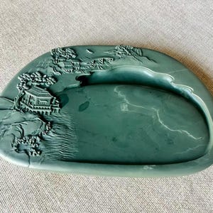 Peut inclure: Une pierre à encre vert jade avec un motif de paysage. La pierre à encre a une forme incurvée avec un puits peu profond et un relief en relief d'une scène de village traditionnel avec des arbres et des bâtiments.