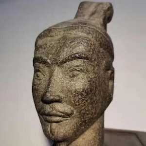 May include: A weathered, gray stone sculpture of a person's head with a detailed face and a textured surface.