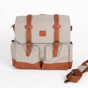 Puede incluir: Bolso mensajero de lona beige con detalles en cuero marrón. El bolso tiene un cierre de solapa con dos correas y hebillas de cuero marrón. Cuenta con dos bolsillos delanteros y una correa para el hombro desmontable.