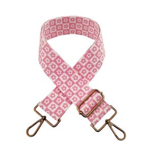 May include: Pink and white floral patterned bag strap with bronze-colored metal hardware. The strap features a repeating flower design and adjustable metal clasps for attaching to a bag. The strap is designed to be worn over the shoulder.