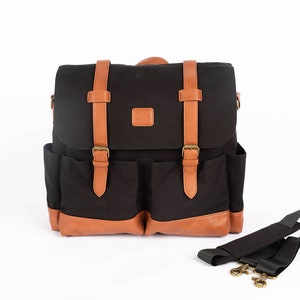 Puede incluir: Mochila de lona negra con detalles en cuero marrón. Cuenta con un cierre de solapa con dos correas de hebilla, dos bolsillos delanteros y una correa para el hombro desmontable. La bolsa tiene forma rectangular y está diseñada para transportar objetos.