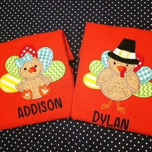 May include: Two red t-shirts with turkey designs. One shirt features a turkey with a bow and the name "Addison". The other shirt has a turkey wearing a pilgrim hat and the name "Dylan". The turkey designs have colorful feathers.