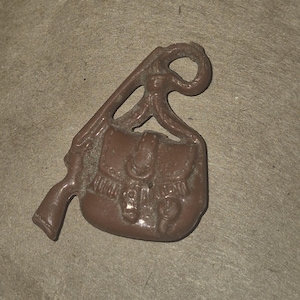 May include: A brown metal charm featuring a rifle and a satchel. The rifle is positioned on the left, with the satchel hanging below and to the right. The charm has a loop at the top for attachment.