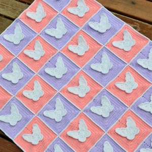 Puede incluir: Una manta de bebé de ganchillo con un patrón de cuadros en tonos de rosa y morado. Cada cuadrado presenta una mariposa blanca de ganchillo.