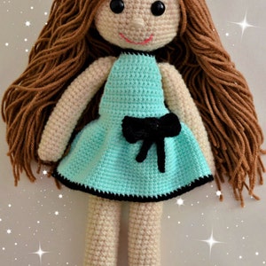 May include: A crocheted doll with long brown hair, wearing a light blue dress with a black bow and black shoes.