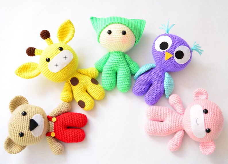 May include: Five crocheted stuffed animals in a circle on a white background. The animals are a yellow giraffe, a green person, a purple owl, a brown bear wearing red overalls, and a pink bear.