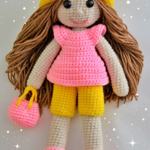 May include: A crocheted doll with brown hair, a yellow hat, a pink dress, yellow shorts, pink shoes, and a pink purse.