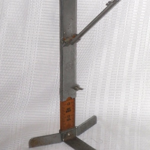 May include: A vintage metal and wood tire tread depth gauge. The gauge has a wooden ruler with inch markings and a metal arm that pivots. The gauge measures up to 24 inches.