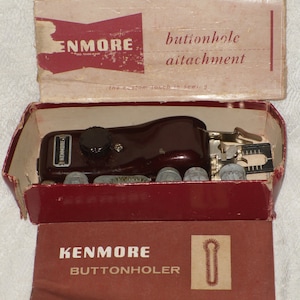 May include: A vintage Kenmore buttonhole attachment in its original box. The attachment is brown and has a silver knob. The box is red and has the words "Kenmore Buttonholer" printed on it.