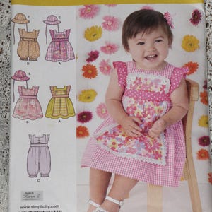 May include: Simplicity sewing pattern 1470A featuring designs for baby dresses, rompers, and hats. The pattern includes various styles with floral and gingham details. A smiling baby girl in a pink dress and white sandals is also pictured.