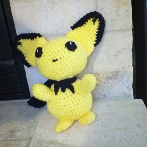 May include: A yellow crocheted plush toy with black ears and a black cape. The toy has a smiling face with black eyes.