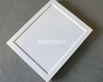 May include: A white, square picture frame with a beveled edge, set at a diagonal angle. The text "stanoyko" is printed in a light grey colour in the centre of the frame. The frame is on a grey surface.