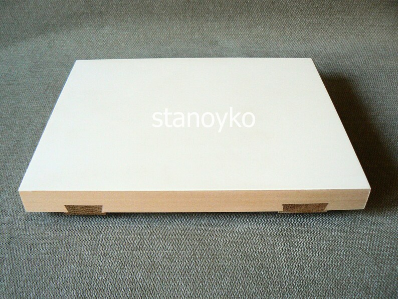 May include: A white rectangular wooden cutting board with rounded edges and a beveled edge. The board has four small wooden feet on the bottom.