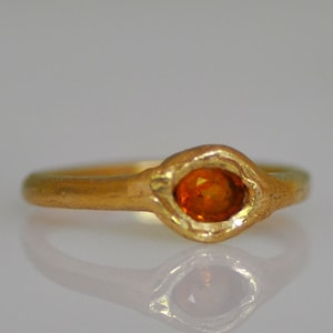 May include: A gold ring with an oval-shaped orange gemstone. The ring has a textured band and a bezel setting that holds the gemstone. The ring is displayed on a reflective surface.