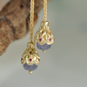 May include: A pair of gold dangle earrings with purple gemstone drops and red accents. The earrings feature a delicate chain design.