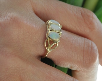 May include: A gold ring featuring three oval-shaped opals set in a row. The band has a delicate, open-work design with a leaf-like pattern on the sides. The opals display iridescent flashes of color, and the metal has a polished, reflective finish.