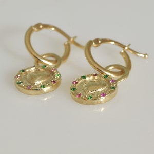 May include: A pair of gold hoop earrings with a heart-shaped charm. The charm is surrounded by a circle of small green and pink gemstones.