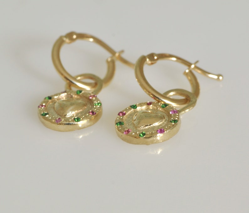 May include: A pair of gold hoop earrings with a heart-shaped charm. The charm is surrounded by a circle of small green and pink gemstones.
