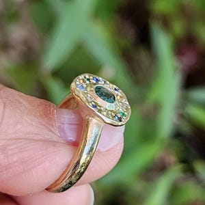 K&ouml;nnte beinhalten: Ein goldener Ring mit einem gro&szlig;en gr&uuml;nen Edelstein, der von kleineren bunten Edelsteinen umgeben ist.