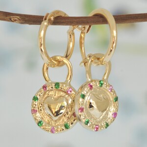 May include: Gold hoop earrings with a heart-shaped charm. The charm is surrounded by a circle of small green and pink gemstones.