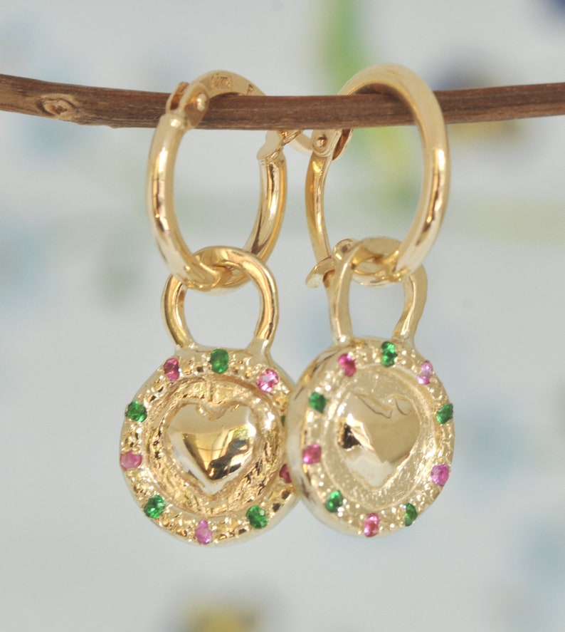 May include: Gold hoop earrings with a heart-shaped charm. The charm is surrounded by a circle of small green and pink gemstones.
